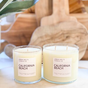 May include: Two glass candle jars with a white label that reads "Urban Melts Candle Co. California Beach Hand Poured in California 11oz | Soy Wax Candle".