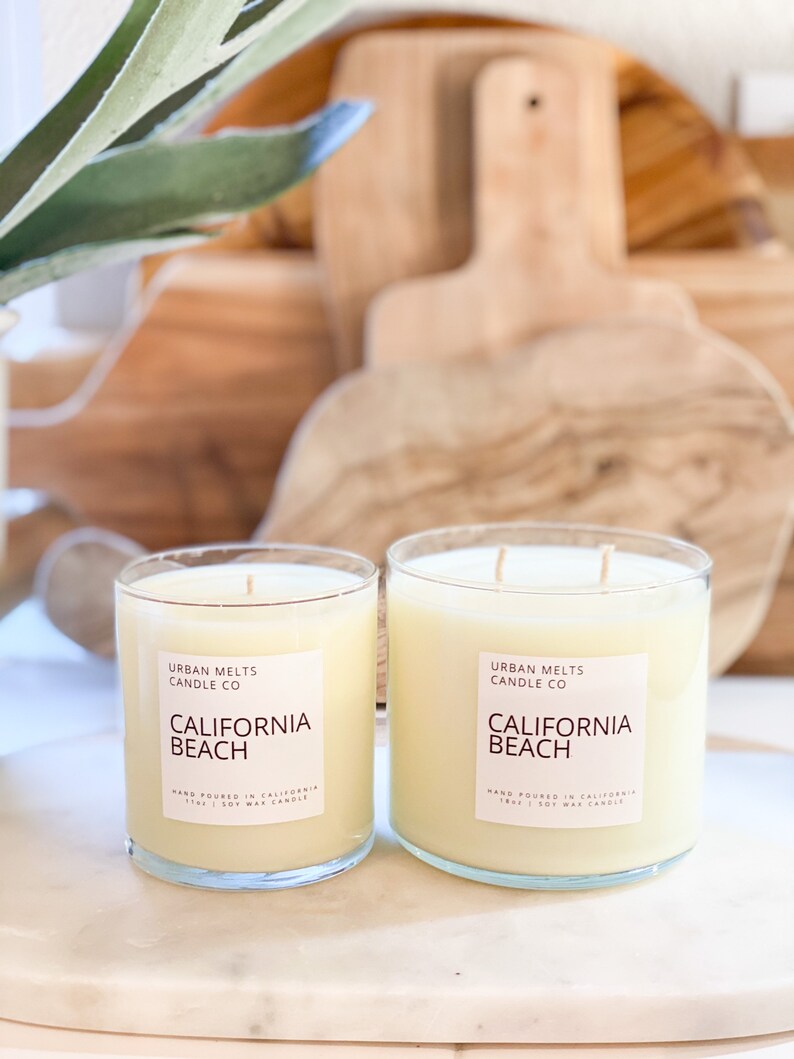 May include: Two glass candle jars with a white label that reads "Urban Melts Candle Co. California Beach Hand Poured in California 11oz | Soy Wax Candle".