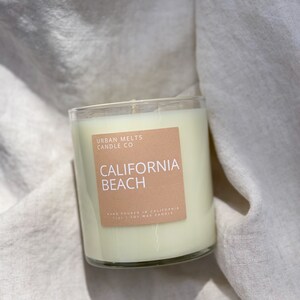 May include: A clear glass candle jar with a beige label that reads "URBAN MELTS CANDLE CO" and "CALIFORNIA BEACH". The label also says "HAND POURED IN CALIFORNIA 11oz | SOY WAX CANDLE".