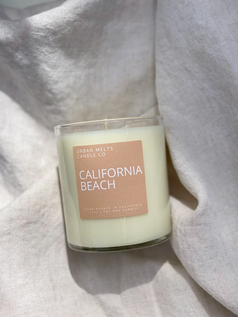 May include: A clear glass candle jar with a beige label that reads "URBAN MELTS CANDLE CO" and "CALIFORNIA BEACH". The label also says "HAND POURED IN CALIFORNIA 11oz | SOY WAX CANDLE".