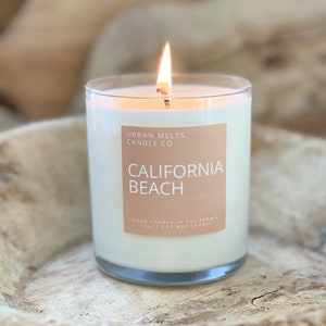 May include: A white candle in a clear glass jar with a label that reads "Urban Melts Candle Co. California Beach Hand Poured in California 11 oz Soy Wax Candle".
