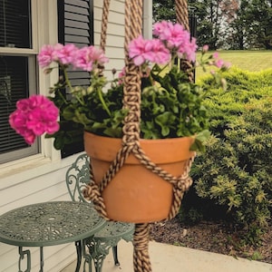 May include: A hanging macrame plant holder with a terracotta pot filled with pink geraniums. The macrame is made of tan and brown rope, and the pot is suspended in front of a white house with black shutters.