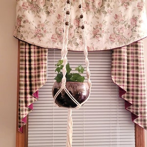 May include: A white macrame plant hanger with wooden beads hangs from the ceiling in front of a window with a floral valance and plaid curtains. A green plant in a brown pot is suspended from the hanger.