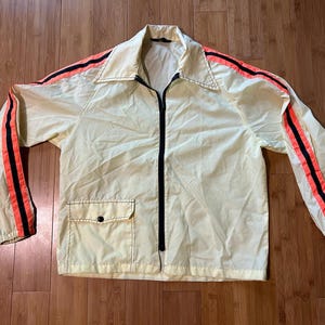 Puede incluir: Chaqueta vintage de color amarillo claro con cremallera negra y un pequeño bolsillo. La chaqueta tiene rayas naranjas y negras en las mangas y en los hombros. Está colocada sobre una superficie de madera.