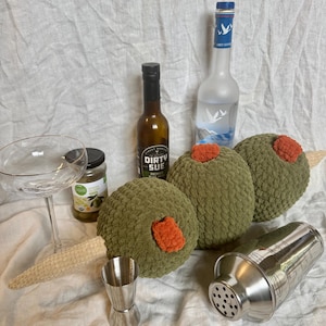 May include: A clear glass coupe, a jar of green olives, a bottle of Dirty Sue olive juice, a bottle of Grey Goose vodka, a stainless steel jigger, a stainless steel cocktail shaker, and a large plush olive garnish on a wooden skewer.