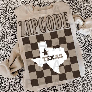 May include: A beige sweatshirt with the text "ZIPCODE" printed on the front. The text is in a brown font with a black outline. Below the text is a brown and white checkered pattern with a white outline of the state of Texas with a star in the center and the word "TEXAS" printed in brown letters.
