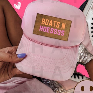以下が含まれることがあります： オレンジとピンクの文字で「BOATS N HOESSSSS」と書かれた茶色の革のパッチが付いたピンクのトラッカーハットです。