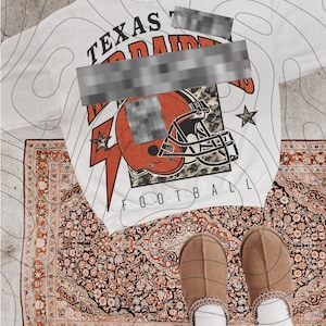 May include: White oversized t-shirt with the text "TEXAS" and "FOOTBALL" with a football helmet graphic. The shirt is laid on a patterned rug with brown slippers and white socks.