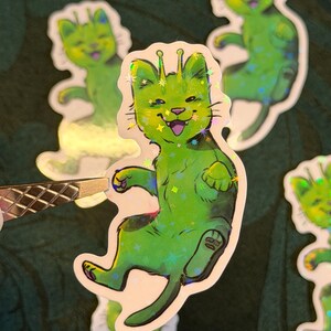 Op de afbeelding: Groene kattenstickers met een holografische afwerking. De stickers tonen een cartoonkat met antennes, een lachend gezicht en steraccenten. Er zijn verschillende stickers zichtbaar, sommige met een metallic glans, op een donkergroene achtergrond.