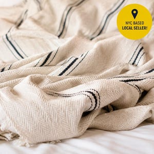 May include: A cream-colored woven throw blanket featuring a subtle herringbone texture and thin, dark navy blue vertical stripes. The blanket has decorative knotted fringe along the edge and a soft, folded appearance with visible fabric weave details.