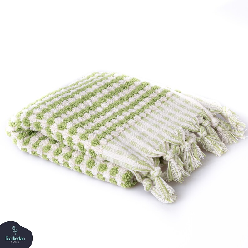 May include: A folded rectangular towel featuring a textured light green and white polka dot pattern on the main body. The edges are finished with a flat woven light green and white checkered border and decorative knotted fringe tassels at the ends.