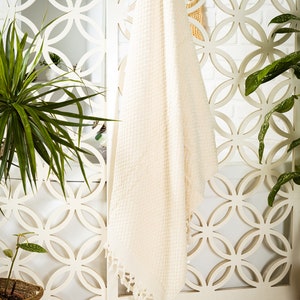 May include: A cream-colored rectangular towel featuring a textured waffle-weave pattern throughout the fabric. The bottom edge of the towel is finished with a row of short, knotted fringe tassels hanging from the hem of the textile.