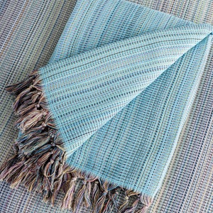 May include: A light blue throw blanket with a fringe border. The blanket is folded in half and has a woven texture.