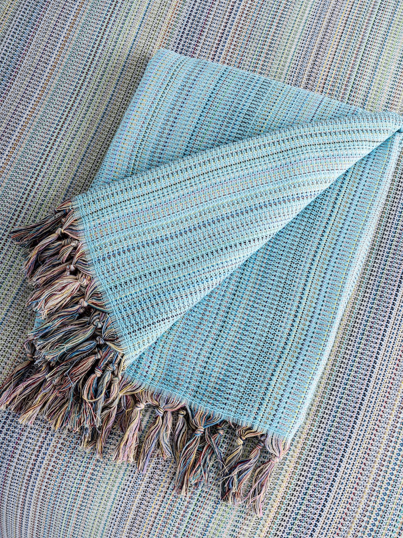 May include: A light blue throw blanket with a fringe border. The blanket is folded in half and has a woven texture.