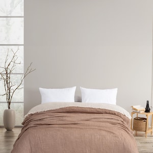 May include: A double-sided bedspread featuring a textured, earthy brown side and a contrasting cream-coloured side. The edges of the bedspread are finished with a frayed, raw fringe detail. Two plain white rectangular pillows rest on the bed.