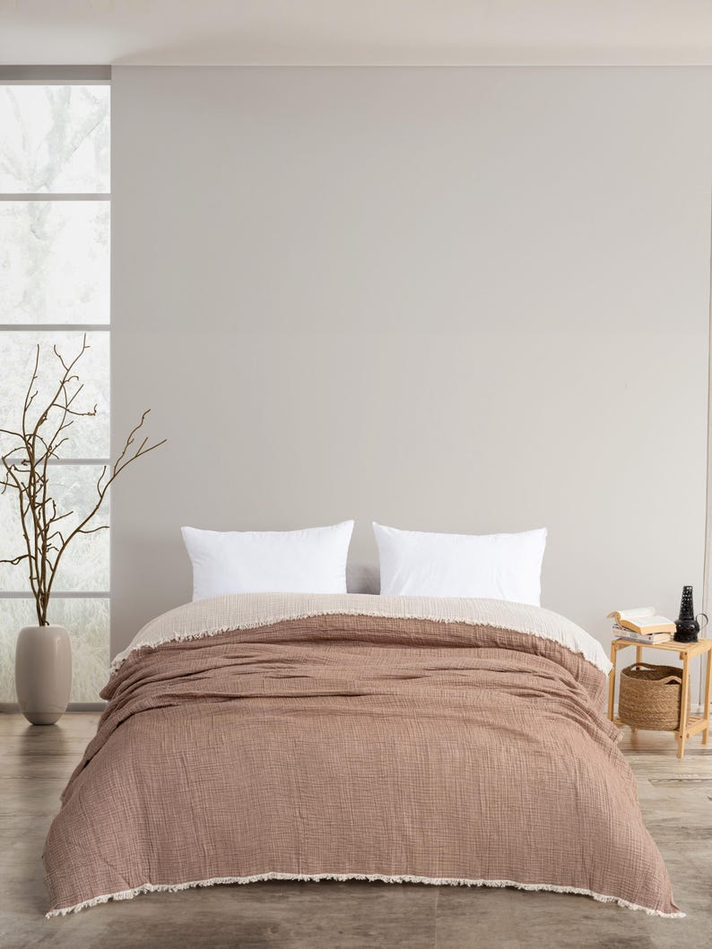 May include: A double-sided bedspread featuring a textured, earthy brown side and a contrasting cream-coloured side. The edges of the bedspread are finished with a frayed, raw fringe detail. Two plain white rectangular pillows rest on the bed.