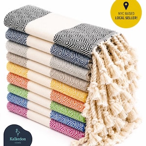 May include: A stack of ten rectangular cotton towels featuring a diamond pattern in black, gray, yellow, orange, green, blue, and pink, separated by white bands. Each towel has twisted fringe ends. A yellow circle contains the text NYC BASED LOCAL SELLER.