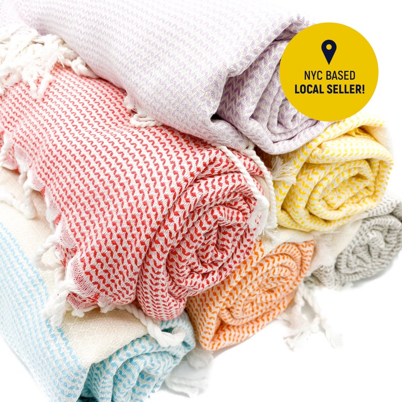 May include: A stack of rolled woven towels in lavender, red, yellow, orange, and light blue, each featuring a white herringbone pattern and fringed ends. A yellow circular badge on the top towel displays the text NYC BASED LOCAL SELLER!