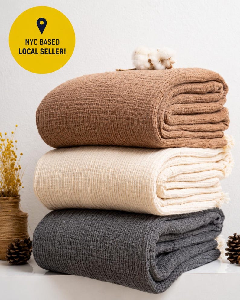 May include: A stack of three folded blankets with a textured, crinkled fabric surface. The top blanket is brown, the middle is cream, and the bottom is dark grey. A small cotton boll sits on the top blanket. A yellow circle contains the text NYC BASED LOCAL SELLER!