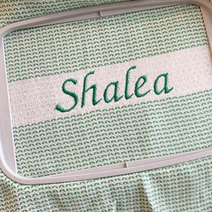 May include: A light green and white woven fabric towel secured in a rectangular embroidery hoop. The fabric features a repeating geometric pattern and a solid white horizontal band with the name Shalea embroidered in dark green cursive script.