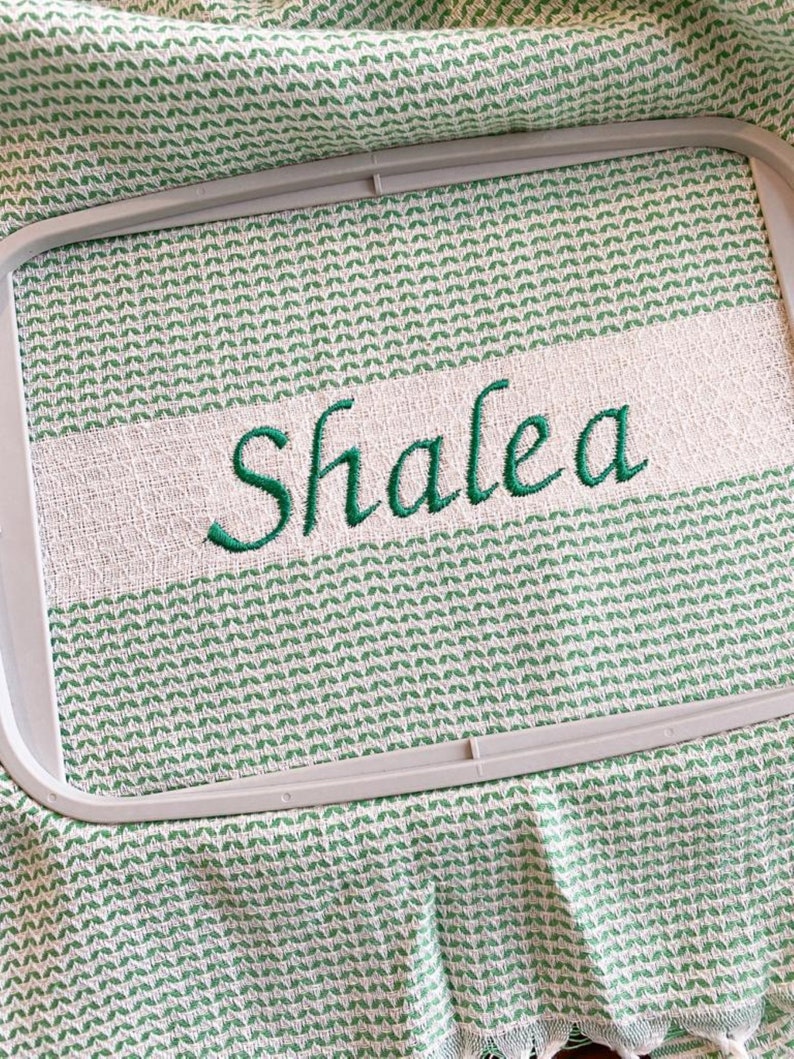 May include: A light green and white woven fabric towel secured in a rectangular embroidery hoop. The fabric features a repeating geometric pattern and a solid white horizontal band with the name Shalea embroidered in dark green cursive script.