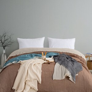 May include: A bed featuring a brown muslin duvet cover with a fringed edge, two white rectangular pillows, and several layered muslin throws in cream, teal, and charcoal grey. A small ceramic mug and an open book rest on the centre of the bed.