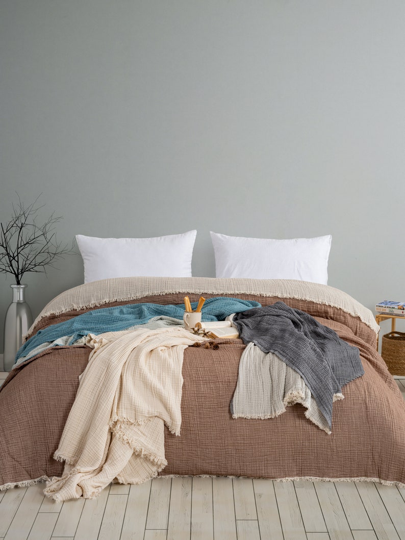 May include: A bed featuring a brown muslin duvet cover with a fringed edge, two white rectangular pillows, and several layered muslin throws in cream, teal, and charcoal grey. A small ceramic mug and an open book rest on the centre of the bed.