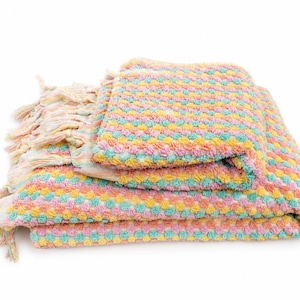 May include: A folded throw blanket featuring a textured, bobble-knit pattern in alternating rows of pastel pink, yellow, and turquoise. The blanket has long, multi-colored fringe tassels along the side edge, creating a soft and tactile appearance.