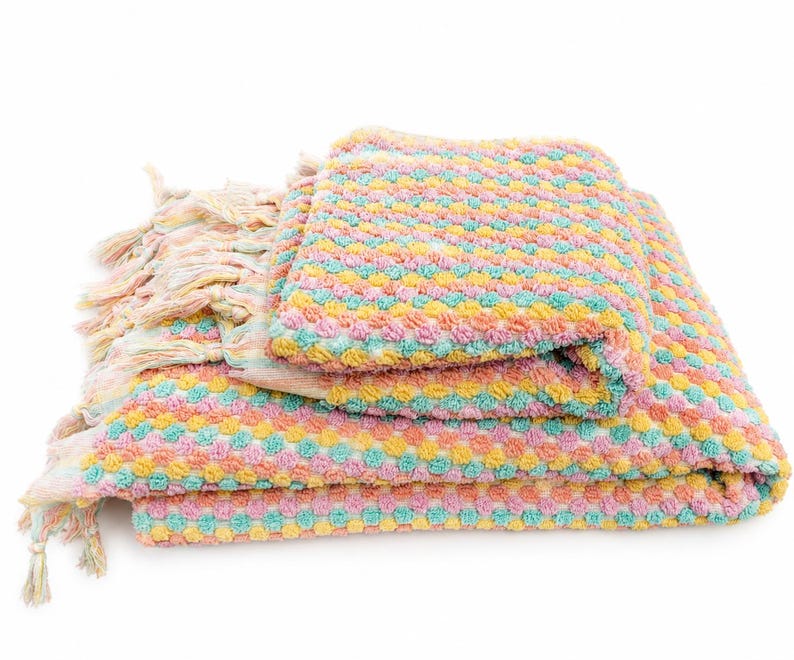 May include: A folded throw blanket featuring a textured, bobble-knit pattern in alternating rows of pastel pink, yellow, and turquoise. The blanket has long, multi-colored fringe tassels along the side edge, creating a soft and tactile appearance.