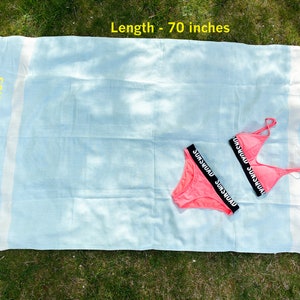 May include: A light blue waffle-weave towel with white stripes and fringed ends, measuring 70 inches in length and 40 inches in width, paired with a bright pink two-piece bikini featuring black elastic bands labeled with the text SUNSQUAD.