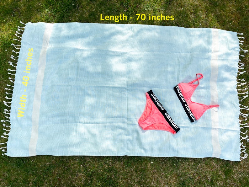 May include: A light blue waffle-weave towel with white stripes and fringed ends, measuring 70 inches in length and 40 inches in width, paired with a bright pink two-piece bikini featuring black elastic bands labeled with the text SUNSQUAD.
