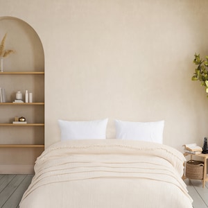 May include: A cream-coloured textured duvet cover with frayed edges draped over a bed. Two plain white rectangular pillows are positioned at the head of the bed. The fabric features a visible grid-like weave pattern throughout the entire surface.