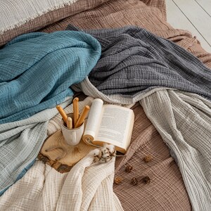May include: A collection of layered muslin blankets in shades of brown, teal, charcoal, and cream. A ceramic mug containing breadsticks, an open book, and small pinecones rest on a circular wooden slice placed atop the textured fabric layers.