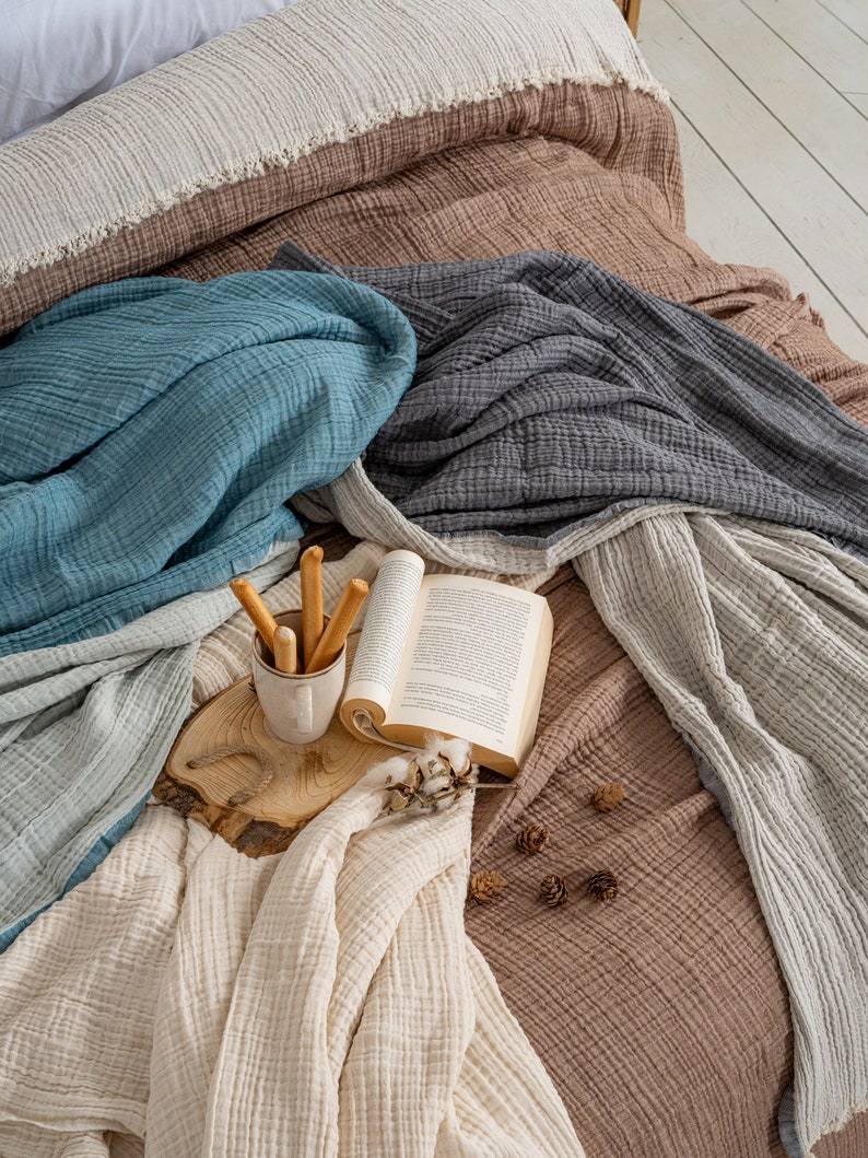 May include: A collection of layered muslin blankets in shades of brown, teal, charcoal, and cream. A ceramic mug containing breadsticks, an open book, and small pinecones rest on a circular wooden slice placed atop the textured fabric layers.
