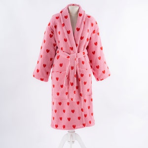 May include: A pink bathrobe with a red heart pattern. The robe has a shawl collar, a tie belt, and two pockets.
