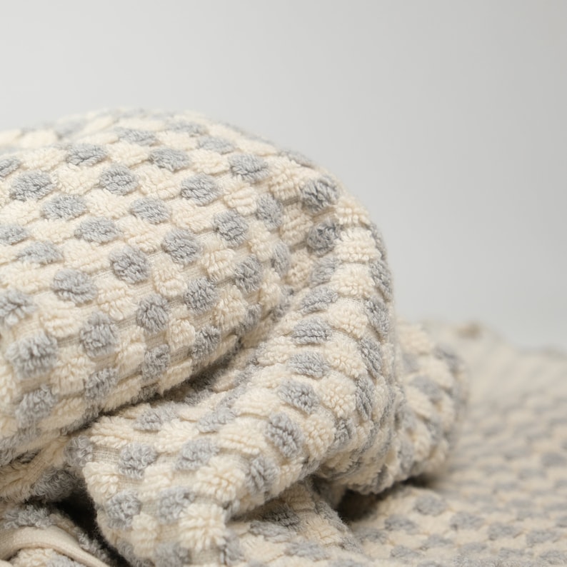 May include: A folded terry cloth towel featuring a textured, repeating pattern of small, raised grey dots against an off-white background. The fabric has a soft, looped pile construction, creating a tactile surface with a consistent color scheme.