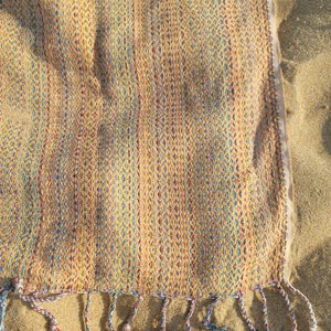 May include: A yellow, blue, and red woven beach towel with fringe on the bottom edge. The towel is lying on a sandy beach.