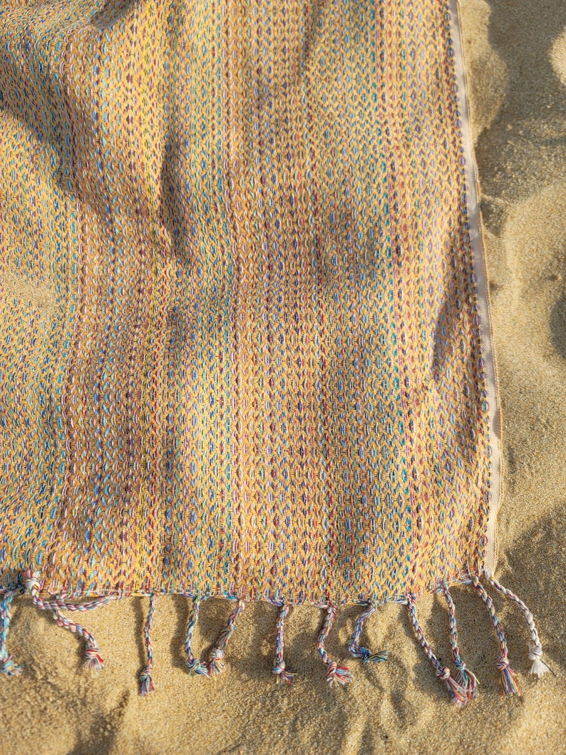 May include: A yellow, blue, and red woven beach towel with fringe on the bottom edge. The towel is lying on a sandy beach.