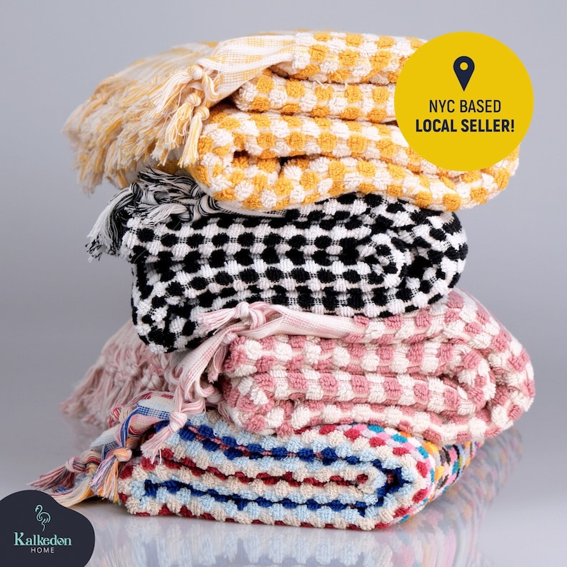 May include: A stack of four folded textured towels with fringed edges. The top towel is yellow and white, the second is black and white, the third is pink and white, and the bottom is multicolored. A yellow circle contains the text NYC BASED LOCAL SELLER!