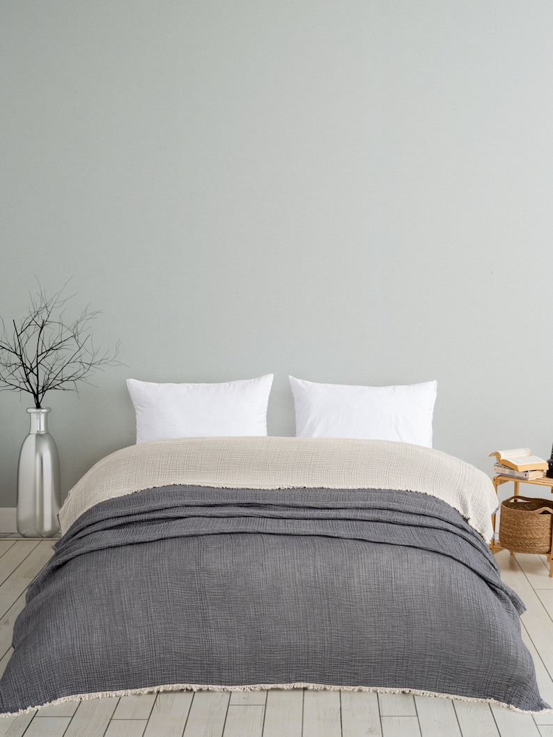May include: A bed featuring a layered bedding set with a dark charcoal grey textured blanket on top of a light beige textured blanket. Two plain white rectangular pillows are positioned at the head of the bed against a light grey wall.