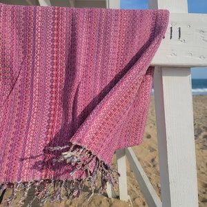 May include: A pink and white patterned beach towel with fringe hanging on a white wooden lifeguard stand.