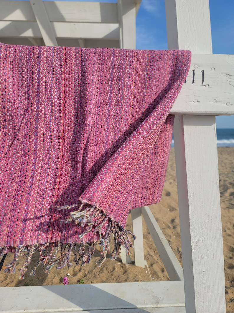 May include: A pink and white patterned beach towel with fringe hanging on a white wooden lifeguard stand.