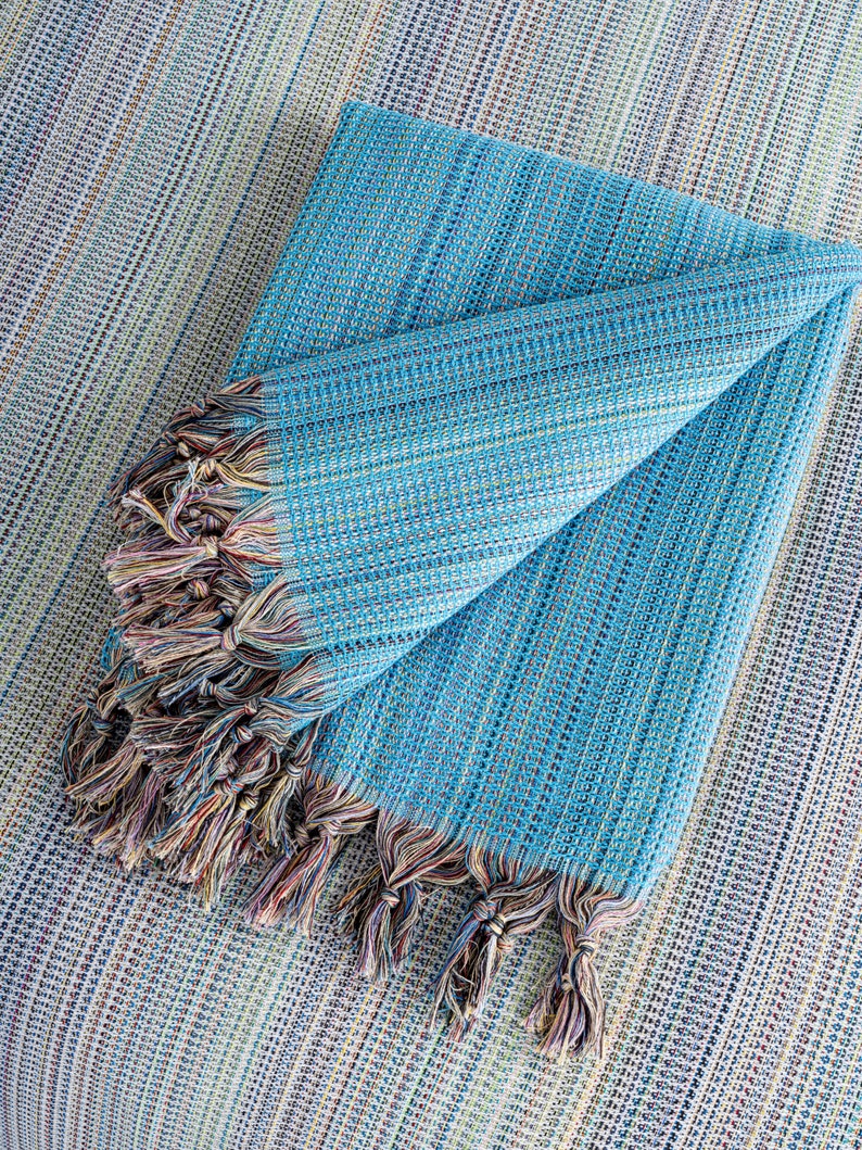 May include: A folded blue and white striped throw blanket with fringe tassels. The blanket is made of a soft, woven fabric.