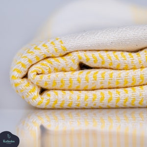 May include: A folded textile featuring a bright yellow and off-white herringbone pattern. The fabric displays a woven texture with visible threads. A small dark logo with the text Kalkedon Home is positioned in the bottom left corner.