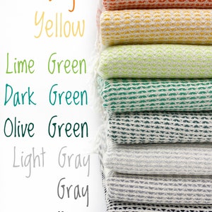 May include: A stack of eight folded woven textiles with fringe, featuring a repeating geometric pattern. The colors from top to bottom are orange, yellow, lime green, dark green, olive green, light gray, gray, and black, with corresponding text labels.