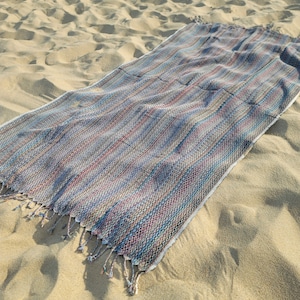 May include: A multi-colored striped beach towel with fringe on the ends. The towel is laid out on a sandy beach.