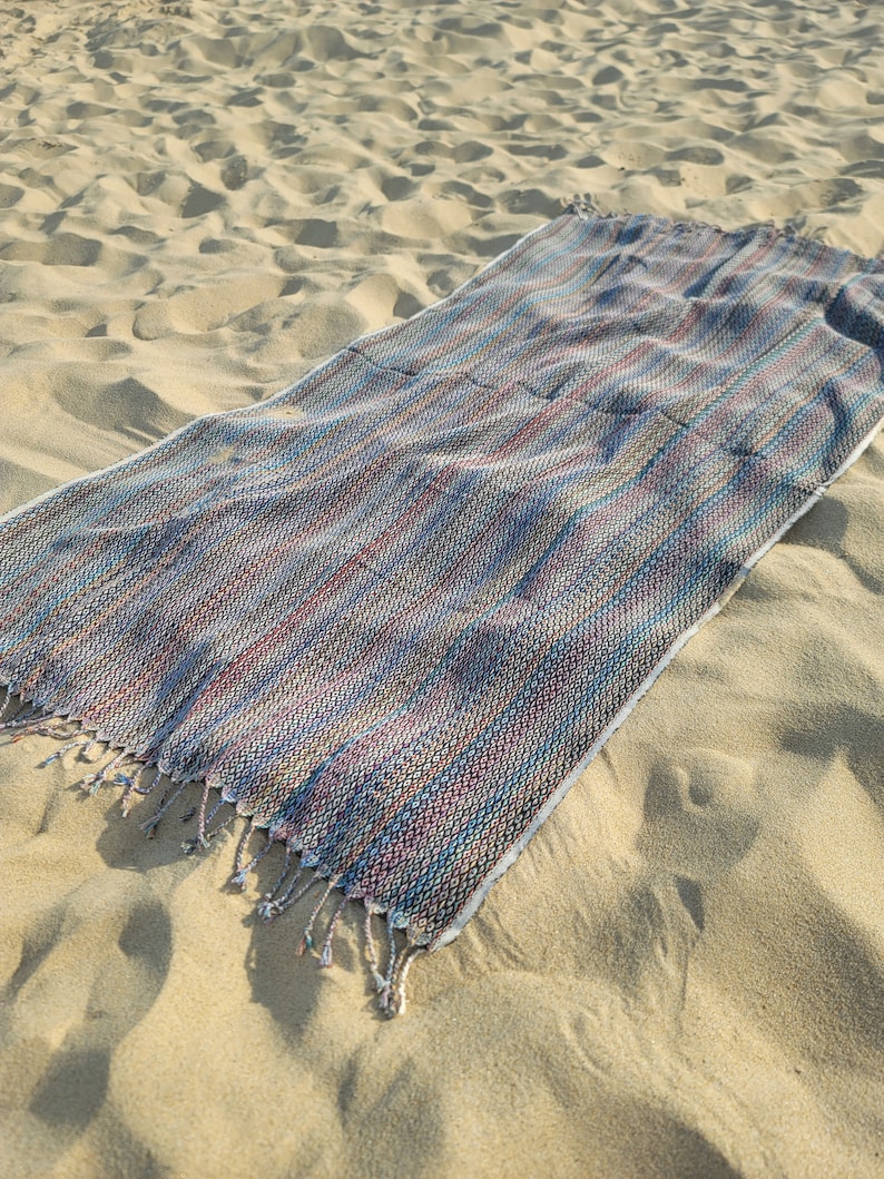 May include: A multi-colored striped beach towel with fringe on the ends. The towel is laid out on a sandy beach.