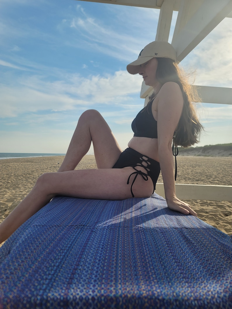 May include: A woman wearing a black bikini top and bottoms with a black lace-up detail on the sides. She is sitting on a blue beach towel with a white hat on her head. The background is a sandy beach with a blue sky and white clouds.