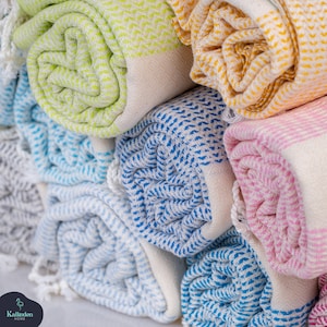 May include: A collection of rolled Turkish towels featuring a woven chevron pattern in lime green, yellow, blue, and pink, each with white fringe ends. The brand name Kalkedon Home is printed in the bottom left corner with a flamingo icon.