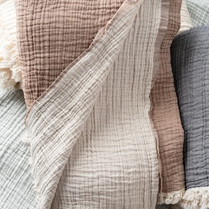 May include: A folded muslin blanket featuring a textured, crinkled fabric in shades of tan and cream. The blanket has a visible grid-like weave pattern and frayed, raw edges along the bottom hem, layered among other similar blankets in various colours.